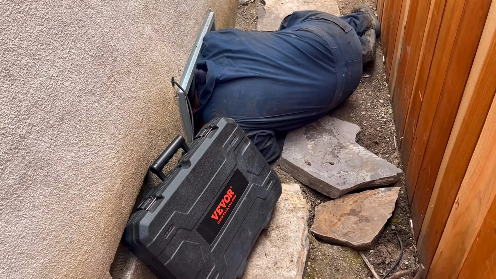 Plumber lying on the ground beside a house wall with a hard case of leak-finding equipment next to him